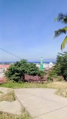 The video shows a panoramic view of a coastal town with buildings, trees, and a mosque with a green minaret. The sky is clear and blue, indicating a sunny day. The foreground consists of a concrete path leading down a grassy slope. The middle ground shows houses with red roofs, lush green trees, and a prominent mosque. In the background, the ocean is visible with some structures on the horizon. The video appears to be taken from an elevated position.