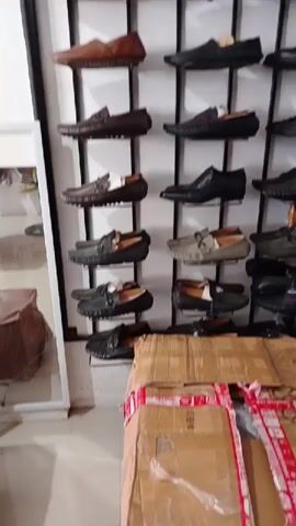The video shows a wall display of shoes, including loafers and sneakers. The shoes are arranged on a black metal rack. The wall is white. In the lower right corner is a cardboard box with red tape. A mirror and small wooden furniture are visible on the left.