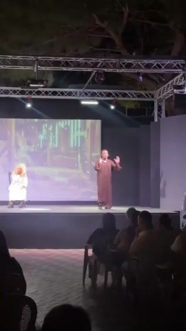 A stage performance is taking place outdoors at night. A man in a brown robe stands center stage, gesturing with his hands as he speaks. To his left, a woman in a white dress sits on a chair, her hands in her lap. A large screen behind them displays an image of a dimly lit, abstract environment. An audience is seated in the foreground, facing the stage. The stage is lit with spotlights.