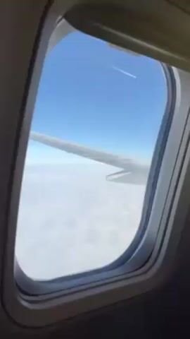 The video shows a view from an airplane window. The wing of the airplane is visible, with white clouds underneath and a clear blue sky above. In the distance, a contrail is visible. The window is clean and the frame is a light gray color.