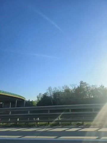 The video shows a highway scene from inside a moving vehicle. The sky is clear and blue, with bright sunlight casting lens flares. On the left, a curved overpass with green railings is visible. A guardrail runs along the side of the highway. In the distance, there are trees and a building with colorful panels. A dark SUV passes by in the right lane.