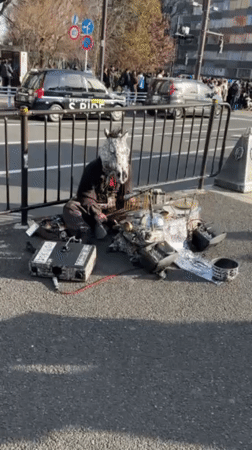Masked performer with electronics sits on Shibuya sidewalk
