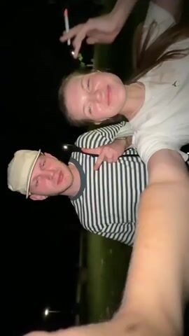 A vertical shot shows a man and a woman posing for a selfie at night. The man is wearing a striped shirt and a baseball cap, while the woman is wearing a white shirt. She holds a cigarette. The man is making a peace sign. They are lying on the grass in a dark outdoor setting. Streetlights are visible in the background. The camera moves around while the woman laughs.