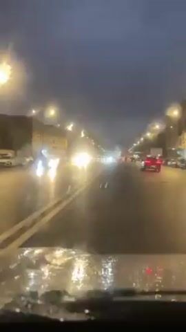 The video shows a driver's point of view driving on a wet city street at night. Streetlights illuminate the multi-lane road, which is reflective from the rain. Other cars are visible, their headlights and taillights blurred by the raindrops on the windshield. A pedestrian is visible crossing the street against the traffic. The sky is overcast and dark.