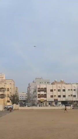 The video shows an urban scene with an airplane flying overhead. The buildings are mainly beige or white with brown window frames and air conditioning units. There are vehicles parked on the streets and a sandy area with a few people moving around. The sky is overcast. A few children yell excitedly as the plane gets closer. The plane is white with red details. It flies directly overhead before continuing into the distance.