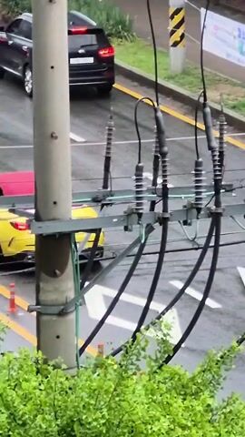 A low-angle, slightly shaky video captures an outdoor scene. In the foreground, a dense cluster of green leaves fills the bottom of the frame. Dominating the mid-ground is a utility pole with multiple wires attached to insulators. To the left of the pole, a bright yellow convertible sports car with its top down is visible, driving away from the camera. The car has a red cover over the rear seats. Orange traffic cones are placed on the wet asphalt road next to the car. A black SUV is parked on the road further back. Behind the yellow car, a white compact car is visible. A person on a motorcycle wearing a helmet and a black jacket with a white backpack is briefly seen riding past the yellow car. The road appears wet, suggesting recent rain. The overall lighting suggests an overcast day.