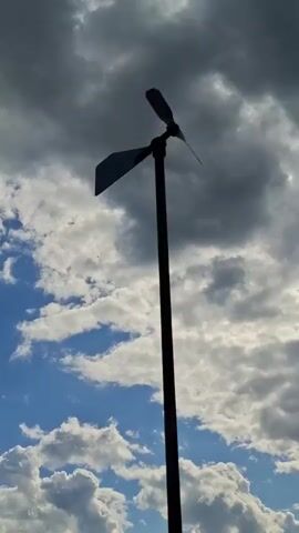 A time-lapse video shows a small wind turbine spinning against a cloudy sky. The turbine is mounted on a pole and has two blades. The sky is blue with some white clouds and darker, stormier clouds in the background. The wind turbine spins slowly at first, then speeds up.