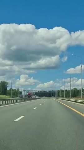 The video shows a view from inside a vehicle driving on a multi-lane highway under a bright blue sky with fluffy white clouds. The highway has clear lane markings and a yellow line along the right edge. On the left side of the highway, there is a barrier and trees. In the distance, there is a large overhead structure, possibly a toll booth or a traffic management gantry, with several vehicles visible underneath it. Some trucks and cars are seen on the highway ahead and to the left. The overall impression is a clear, sunny day on a highway.