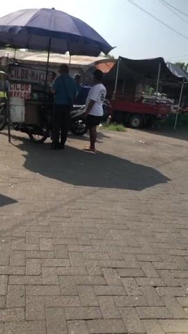 A street vendor is selling food from a cart. The cart is mobile and appears to be attached to a motorcycle. The vendor is standing behind the cart, serving a customer. The customer is standing in front of the cart, receiving their order. The scene is outdoors, and the weather appears to be sunny. There are other stalls and vehicles in the background, suggesting a busy market or street. The vendor's cart has a sign that says "CILOR MAKLOR."