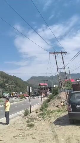 The video opens with a view of a road and a mountainous landscape under a clear blue sky with scattered clouds. Two men are visible on the side of the road, one wearing a blue t-shirt and the other a yellow shirt. The camera then pans to the right, showing several buildings nestled on the slopes of the mountains. These buildings appear to be a mix of residential and commercial properties, with some featuring red roofs. As the video continues, the camera moves further along the road, revealing more of the rural, mountainous environment. The road is paved and appears to be a main thoroughfare. There are several cars parked along the roadside and some in the distance. The terrain is characterized by lush green hills and trees. The video concludes with a shot of the road stretching into the distance, with more buildings and construction visible on the mountainside.