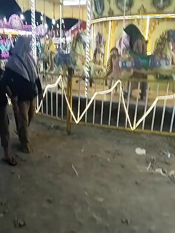 The video depicts an outdoor amusement park or carnival scene at night. The main focus is a brightly lit carousel with yellow and purple lights, where several people, including children and adults (some wearing hijabs), are enjoying rides on the horse figures. In the foreground, a young boy in a black shirt and a woman in a gray hijab are walking past the carousel. The ground is unpaved, appearing to be dirt or gravel, with some litter visible. As the camera pans, more of the carnival atmosphere is revealed, with various other amusement rides adorned with colorful lights (blue, green, red, purple) in the background. Several people are seen walking around, standing, or sitting near tents and stalls. A man in a plaid shirt walks away from the camera, and another woman in a light blue hijab is seen standing near a tent on the left side of the frame. The scene is illuminated by the many lights of the rides and stalls, indicating it is nighttime. No explicit text is visible in the video. The setting suggests a festive local fair or market in Sragen, Indonesia.