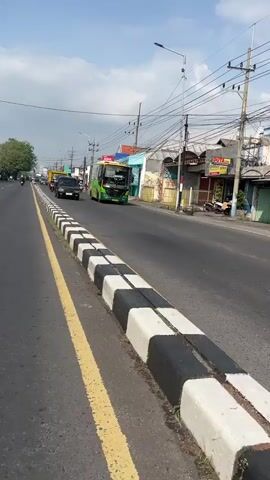 The video shows a street scene with a green bus and a black SUV driving on the road. The street is lined with a sidewalk and buildings on the right side of the frame. There are utility poles with wires visible overhead. The sky is partly cloudy. The road has yellow and white lane markings. The bus is in the left lane and the SUV is in the right lane, both moving forward. The camera appears to be mounted on a moving vehicle, as indicated by the motion blur and the perspective of the road.