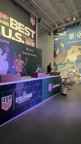 A man walks across a large room with a blue mural of people and soccer balls on the wall behind him. The mural also features the USA soccer logo and a Volkswagen logo. In front of the mural, a large counter with a blue and green checkered design is visible. The counter has the USA soccer logo and "Chobani" branding. On the counter, there are two wooden boxes and a soccer ball. To the left of the counter, a large black banner with white text that says "BEST U.S." is hanging from the ceiling. To the right of the counter, a green lift is parked. The floor is a dark gray concrete. The lighting in the room is artificial and appears to be coming from overhead.