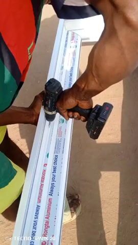 A person is securing a corner of a white aluminum window frame with a drill. They are outside on bare ground. The person wears a multi-colored shirt and yellow shorts, as well as sandals. The text on the aluminum window frame includes "Kongtai Aluminum, Always your best choice." A "Tecno Spark 20" watermark is at the bottom left of the frame.