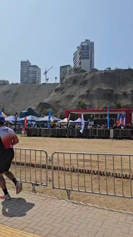 The video shows a crowd of people gathered behind a metal barrier, likely to watch an event taking place on a sandy area.  There are tents, flags, and a red structure behind the crowd. In the background, there's a hill with buildings and palm trees on top, against a clear blue sky. A drone is seen flying overhead.