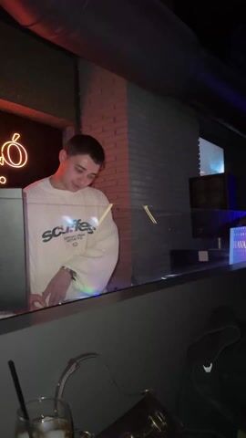 The video shows the inside of a club. The video starts with a shot of a DJ at a booth, then pans across the room, showing people dancing and having fun. A woman with long black hair briefly covers her face with her hands and then smiles. The room is dimly lit with blue lights. There are tables, chairs, and drinks visible. The walls are brick and painted blue. The club is crowded.