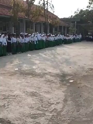 The video shows a group of children lined up in rows on a concrete outdoor space, appearing to be a schoolyard. They are dressed in white shirts and green skirts or pants, indicating a school uniform. Most of the girls have white headscarves. The children stand facing toward the right of the frame. A building, likely the school, is visible in the background. There are trees and some smaller groups of students seated or standing further to the right of the main group. The scene seems to capture a school assembly or some formal event. The video camera is handheld, with some shaking evident.