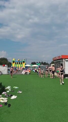 A crowded outdoor water park scene unfolds under a bright, cloudy sky.  In the foreground, a lush green artificial turf area is scattered with numerous pairs of footwear, including sandals, flip-flops, and sneakers. A set of green steps covered in the same turf leads up to a higher level. A woman in a black and white bikini is walking down these steps, followed by two young children. One child, a girl in a bikini and a pink inflatable ring, is descending the steps. Another child, a boy in green shorts, is at the bottom of the steps. To the right, several adults and children are present, many in swimwear. Some are standing, while others are seated on lounge chairs. In the mid-ground, a large inflatable water slide structure with yellow and green sections is visible, with a line of people waiting at its base. In the background, trees and some structures, including what appears to be a building with orange and white signage, are present. Several other inflatable structures and white tents are scattered throughout the area. The overall impression is a busy recreational environment.