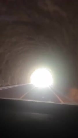 The video shows the interior of a vehicle traveling through a tunnel. The camera is positioned from the driver's perspective, looking forward. Initially, the tunnel is dark with rough, textured walls, and a bright light is visible at the tunnel's exit. As the vehicle moves forward, the tunnel opens up to a bright, overcast sky. The road ahead is paved with white lane markings and is surrounded by green foliage and trees. The scene suggests a journey from a dark enclosed space into an open, natural landscape.