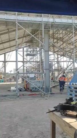 The video shows a construction site with scaffolding. In the background, a person in an orange vest and hard hat walks across the scene. In the foreground, there are stacks of materials and a wooden table with various items on it. The environment appears to be an indoor construction area with a large roof structure.