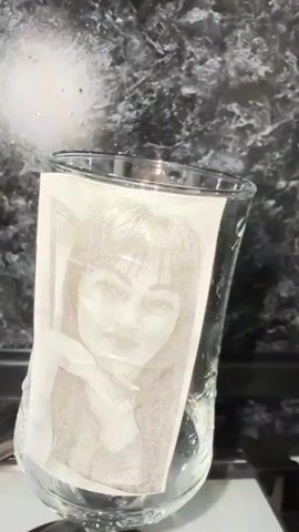 A black and white image of a woman's face is taped to the inside of a clear glass that contains water. The glass is tilted and appears to be a wine glass with a stem. The background is dark and textured, possibly a countertop or backsplash. The lighting suggests it is indoors.