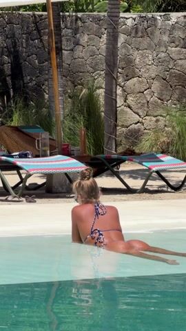 A woman in a blue and white patterned bikini lies in a pool of clear, light blue water. She is seen from behind with her hair in a bun. She wears sunglasses. Her body is submerged from the waist down. The pool is surrounded by sand and two colorful lounge chairs. Behind the chairs is a stone wall and some greenery. There is a glass of liquid on a small table next to one of the chairs.
