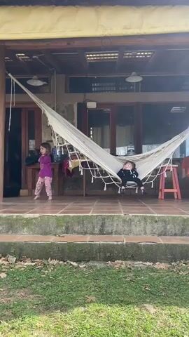 Two young girls are visible on a covered porch. The girl on the left, wearing a purple long-sleeved shirt and patterned leggings, is standing and appears to be speaking. The girl on the right is lying in a white hammock, wearing a black long-sleeved shirt and dark pants. The porch has a tiled floor and is furnished with wooden tables and chairs. In the background, glass doors lead into a building. A grassy area with fallen leaves is in the foreground. The girl on the left then walks towards the right, then turns back and approaches a table. The girl in the hammock is propped up and appears to be looking at something in her hands. The girl on the left then moves to the right and bends over a table. The girl in the hammock is still in the hammock. The girl on the left is still bending over a table. The girl in the hammock appears to be holding something pink in her hands.