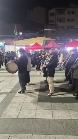 The video captures a group of people playing wind instruments, likely flutes, in an outdoor setting at night. They are arranged in a line, facing the same direction. The people are dressed in casual clothing, and some are wearing hats. The instruments are long and made of wood, with colorful bands near the top. In the background, there are red tents, buildings, and lights, suggesting a market or street fair. The ground is paved with square tiles. The camera is positioned at a slightly elevated angle, looking down at the musicians.