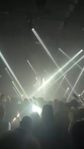 The video shows a crowd of people in a dark, smoky environment, likely a nightclub or concert venue. Bright white spotlights shine down from above, creating a dramatic lighting effect. The crowd appears to be enjoying themselves, moving and dancing to the music. Some people are holding up their phones, presumably to record the event. The camera angle is from within the crowd, offering a view of the stage area, though the performer is not clearly visible due to the lighting and distance.