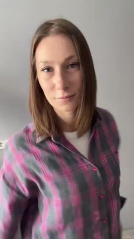 A young woman with shoulder-length brown hair and light makeup is standing against a plain gray wall. She is wearing a pink and gray plaid flannel shirt over a white t-shirt. In the background, on the left side of the frame, there is a white electrical outlet with three sockets. The woman smiles at the camera, turns her body to the side to show her back, then turns back to face the camera and claps her hands. She then turns her head to the side and looks over her shoulder before returning her gaze to the camera. Her movements are fluid and graceful.