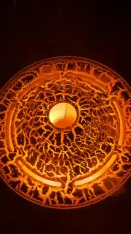 The video features a close-up shot of a circular, decorative object, possibly a lamp or ceiling fixture, illuminated with a bright orange light. The surface of the object is textured with a pattern resembling cracked earth or lava flows. In the center, there is a smooth, spherical bulb emitting a warm, yellow glow. The background is dark and out of focus, highlighting the illuminated object. A voice speaking in an unfamiliar language can be heard.