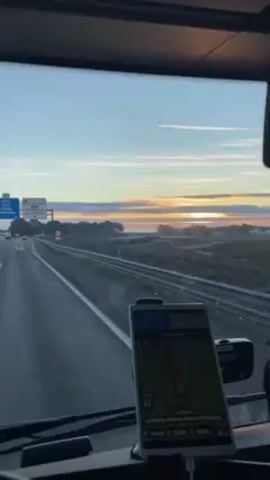 The view from inside a vehicle driving on a highway reveals an open horizon with a rising sun. The road ahead is clear with minimal traffic. Road signs indicate exits for "AP-2" towards "Fraga, Lleida, Barcelona" and "A-110" towards "Bujaraloz, La Almolda." A GPS device mounted on the dashboard displays a map and navigational information. The vehicle maintains a steady pace, moving forward on the highway as dawn breaks.