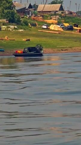 A hovercraft is docked on the bank of a body of water, with a small village visible in the background. The village consists of several houses, some appearing to be made of wood, situated on a grassy slope. Various objects are scattered around the village, including what appear to be cars, logs, and other debris. The water in the foreground is rippling, suggesting a slight breeze or movement. The sky is overcast.
