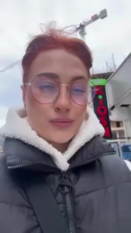 A woman with reddish-orange hair, wearing round-framed glasses and a dark gray puffer vest with a white fleece lining, stands outside in what appears to be a windy urban environment. In the background, there is a tall construction crane and an unfinished building. A sign reading "KIOSK" is visible to the side. The woman is facing the camera with a neutral expression. The wind is blowing her hair.