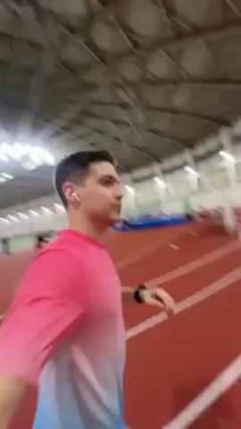 A man is walking on an indoor running track. He is wearing a short-sleeved shirt that is pink on top and fades to blue on the bottom, along with black shorts. He is holding the camera in front of him, looking at the camera as he walks. The track is red with white lines marking the lanes. There are other people on the track in the distance, and some equipment like hurdles are visible. The building appears to be a large indoor sports facility with a high ceiling and bright overhead lights.