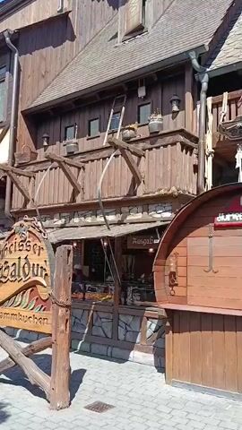 The video pans across an outdoor area with a roller coaster track visible in the background. Several wooden structures, resembling a medieval village, line the area. A sign with German text reads "Heisser Baldu Flammenkuchen". People are seen standing at barrels, some gathered in conversation, while others are seated at tables under an umbrella. The ground is paved with cobblestones. The scene appears to be a theme park or outdoor dining area on a sunny day.