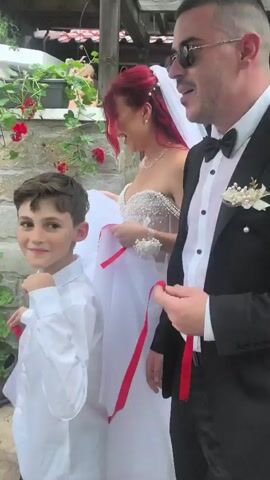 A video shows a wedding celebration outdoors. A groom in a black suit with a white boutonnière and sunglasses adjusts his glasses. A bride with red hair wears a white strapless gown with pearl detailing and a veil. A young boy in a white shirt and black bow tie stands beside the groom. The groom holds several red ribbons tied to a white fabric. Another man in a light green suit holds a red banner with white writing and fringe. Other guests are visible in the background, some holding drinks. The celebration takes place under a white pergola adorned with greenery and red flowers.