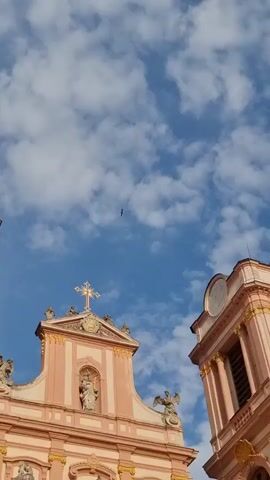 The video is a vertical shot of the facade of a large, ornate building, likely a church or monastery, with a blue sky and scattered clouds in the background. The building is predominantly pink and cream with gold accents. Two people are visible walking up a wide set of stone stairs leading to the entrance. One person is wearing a white outfit and the other is wearing dark clothing. The facade features columns, statues, a large cross at the peak, and architectural details. A bird is also visible flying in the sky.