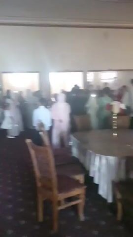 The video shows an indoor event with several people present. The room has a patterned carpeted floor and is furnished with tables and chairs. In the background, there is a stage area with a backdrop that appears to be decorated with lights. A band or musicians are visible on the stage, with at least one person playing a drum and another holding a stringed instrument. Some attendees are seated at tables, while others are standing and mingling. There is a second level or balcony overlooking the main floor. The lighting in the room is mixed, with some bright spotlights and ambient lighting. People are dressed in various attire, some in traditional clothing. The overall atmosphere suggests a celebration or gathering.