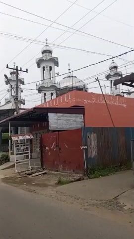 A white mosque with a silver dome and a tall minaret stands in the background. In the foreground, a red building with a corrugated metal roof is visible, along with a white structure that appears to be a small stall or kiosk. A grey pickup truck is parked on the left side of the road, and a black motorcycle is parked in front of it. The road is paved and appears to be wet, suggesting recent rain. The sky is overcast. The video is filmed from a moving vehicle, giving a dynamic perspective of the scene. The overall impression is that of a street view in a residential or commercial area, with the mosque being a prominent landmark.
