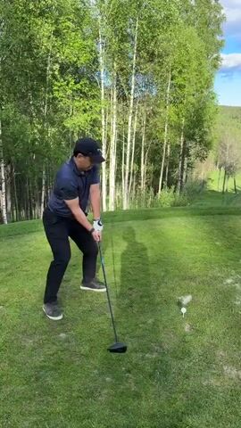 The video depicts a man playing golf on a green golf course. The man is wearing a navy blue polo shirt, black pants, and a black baseball cap. He swings a golf club, and the ball is visible flying through the air. The course is surrounded by trees and hills with a blue sky and scattered clouds. After swinging, the man pauses, checks where the ball landed and then looks up toward the sky. A yellow tee is visible on the ground near where the golf ball was positioned.