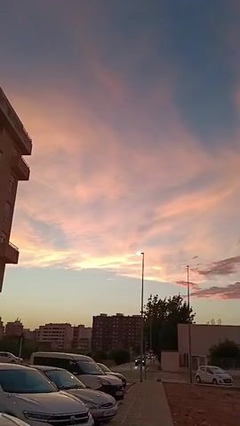 The video shows a time-lapse of a sunset over an urban environment. In the foreground, there is a sidewalk with parked cars. Beyond the sidewalk, there are buildings and trees. The sky is filled with clouds that are illuminated by the setting sun, creating a beautiful display of pink and orange hues. The camera pans slightly to the left throughout the video, showing a wider view of the sky and the cityscape. There are no people visible in the video.