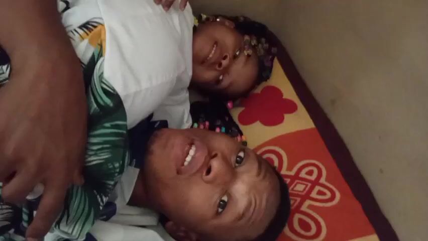 A video shows a man and a child lying on a patterned rug. The man is in the foreground, wearing a blue and white patterned shirt with a green leafy design. The child is behind him, wearing a white shirt. Both are smiling and looking at the camera. The man opens his mouth wide and sticks his tongue out. The child reaches out and touches the man's face. The background is a plain, light brown wall.