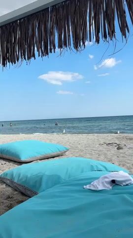 The video depicts a serene beach scene under a clear blue sky with scattered clouds. The foreground features two large, turquoise beanbag chairs placed on a sandy beach. A white t-shirt is draped over the one closer to the camera.

In the background, the calm, turquoise sea stretches towards the horizon. Several people are visible in the water, some swimming or wading. A seagull stands on the wet sand near the water's edge, looking towards the camera. Another seagull is visible further out in the water. The scene is shaded by a thatched roof structure, suggesting a beach lounge area.

The overall atmosphere is one of relaxation and summer vacation. The bright sunlight and clear weather indicate it is daytime, likely midday or early afternoon. The location is identified as Paralia Mesis, Greece, which is a coastal area. The gentle waves and the presence of people enjoying the water suggest a warm and pleasant day for beach activities. There is no visible text in the video.