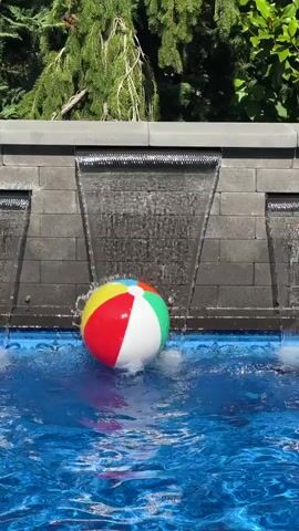 A colorful inflatable beach ball floats in a blue swimming pool. Water cascades down from a textured gray wall behind the pool, creating waterfalls. The beach ball is predominantly yellow, red, and blue, with some green and white sections. It appears to be bobbing in the water, likely due to the water flow or ripples. The background shows lush green foliage.