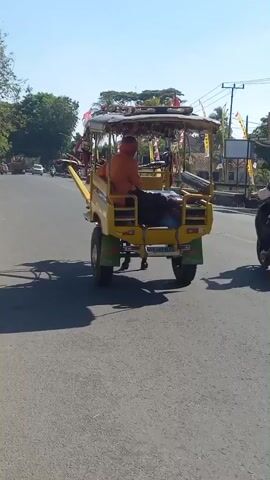 A yellow motorized rickshaw is driving down a paved road on a sunny day. The rickshaw has a canopy and is decorated with small flags. A person wearing an orange shirt is driving the rickshaw. There are other vehicles and buildings visible in the background.