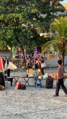 The video shows a beach scene with several people, stalls, and flags. In the foreground, a person is walking on the sand. To the left, there are flags, including the Palestinian flag and the Malaysian flag, planted in the sand. There are also bicycles and a scooter. In the middle ground, there are several stalls set up with tables displaying various items, possibly for sale. People are seen interacting at these stalls, and some appear to be selling or buying goods. Bubbles are being blown in the air, adding to the lively atmosphere. In the background, there are trees, buildings, and more people walking along the beach. The overall environment suggests a marketplace or a gathering of people enjoying a day at the beach. The lighting indicates it might be late afternoon or early evening.