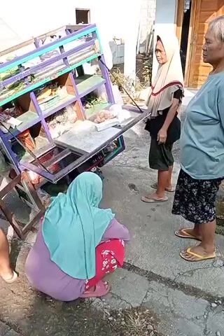 A vendor cart with multiple shelves, laden with various food items, has tipped over. Three women are present. One woman, wearing a light blue hijab and red patterned leggings, is squatting near the tipped cart, seemingly attending to the spilled items. Another woman, wearing a beige hijab and dark shorts, stands upright, observing the scene. The third woman, wearing a light blue t-shirt and a dark patterned skirt, stands further to the right, also observing. The cart appears to be on a paved surface, possibly a street or walkway, with a building visible in the background. The lighting suggests it is daytime.