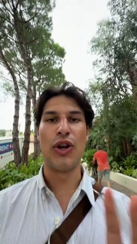 This media file captures a man filming a video of himself as he walks towards a large, luxurious house. The house appears to be a villa, with a sprawling facade, a fountain in the courtyard, and lush greenery surrounding it. Two black vans are parked in front of the house, and several people are visible in the background, including a person on a bicycle and a couple walking near the fountain. The man speaking is wearing a light-colored, striped shirt and a brown strap across his shoulder. He has dark hair and appears to be smiling and excited, suggesting a positive emotional state. The overall scene conveys a sense of affluence and a grand arrival. The time of day seems to be overcast, possibly late afternoon, given the diffused lighting. There is some visible text on a white sign in the background that reads "RENT + CARE". The setting suggests a high-end resort or a private estate in Vallauris, France. The activity is the man documenting his arrival and initial impressions of the location.
