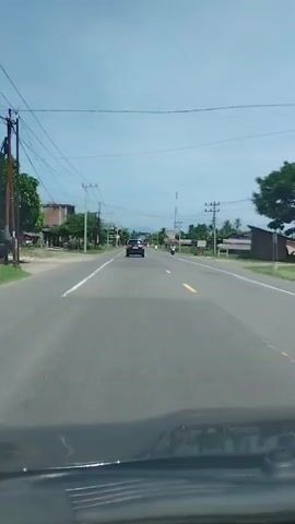 The video shows a view from inside a car, looking out at a straight, two-lane road. A dark-colored car is driving ahead in the same lane, further down the road. To the right of the car, a person is riding a scooter in the same direction. Power lines run along both sides of the road, supported by utility poles. Buildings and trees are visible on either side of the road, set back from the pavement. The sky is clear and blue.